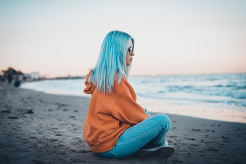 Young hipster woman in orange sweatshirt with blue hair and glasses sitting on sea beach