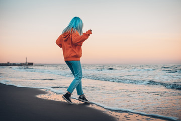 Young hipster woman in orange sweatshirt with blue hair playing with sea waves on beach