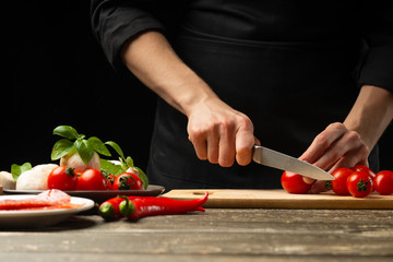 The chef cuts cherry tomatoes with vegetables. For cooking pizza, tomato sauce, salad. A delicious meal concept, cooking. On a black background for design or lettering text. Close-up