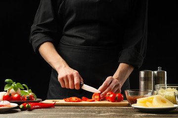 The chef cuts cherry tomatoes with vegetables. For the preparation of pizza, tomato sauce, salad. A delicious meal concept, cooking. On a black background for design or lettering text.