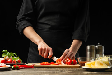 The chef cuts cherry tomatoes with vegetables. For the preparation of pizza, tomato sauce, salad. A delicious meal concept, cooking. On a black background for design or lettering text.