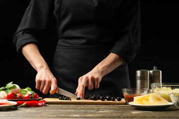 The chef cuts the olives. For the preparation of pizza, salad. A delicious meal concept. On a black background for design or lettering text.