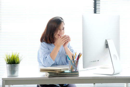 Young asian woman holding a coffee cup with smiling face while working with computer, positive emotion at working desk background, casual office life, working at home concept