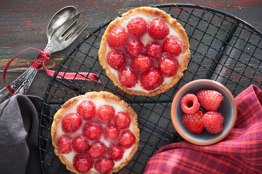 Red raspberry shortbread tarts with vanille custard and glazed fresh raspberries on cooling rack on dark brown wood - Powered by Adobe