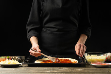 Professional chef prepares Italian pizza, spreads tomato sauce. On a black background, horizontal...