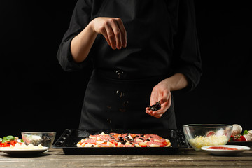 Chef sprinkles pizza with olives. Frost on the move. A concept of delicious food and healthy food. On a black background for design or lettering text.