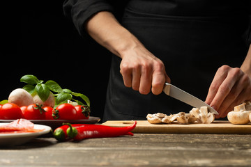 The chef cuts champignon mushrooms with vegetables. For cooking pizza, mushroom sauce, salad. A delicious food concept, cooking. On a black background for design or text writing. Close-up