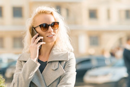 Outdoor portrait of young blonde woman with curly hair smiling and talking on a mobile phone on sunny autumn day in the city
