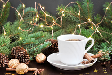 Obraz na płótnie Canvas Winter season holidays mood. Cup of coffee with cinnamon surrounded by fir branches and cones with lights garland on rustic wooden background. flat lay