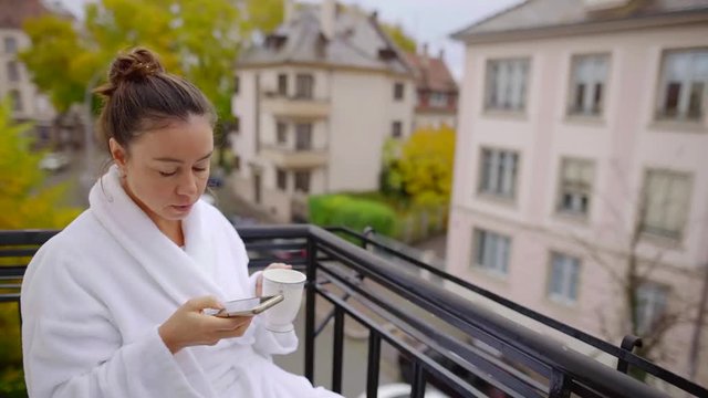 Portrait of a brunette woman with a cup of coffee and smartphone on balcony wearing white robe.