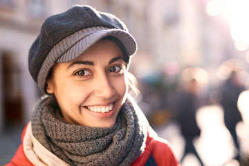 Portrait of a young smiling woman in red jacket,gray scarf and cap. Woman posing outdoors on the street at sunny autumn or spring day