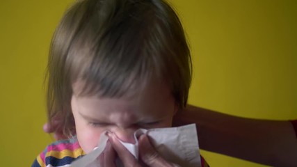 Adult helping little girl to blow nose by napkin indoor, face closup shot. Woman keeping napkin near her daughter face while toddler blowing her nose. Mother helps baby taking care during sickness
