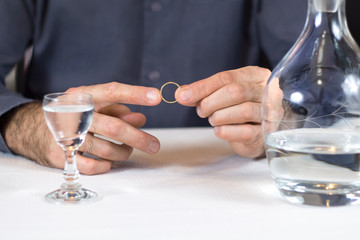 Problemy w małżeństwie. Mężczyzna trzyma obrączkę ślubną w dłoniach. Obok na stole stoi butelka z alkoholem oraz napełniony kieliszek do wódki.