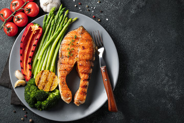 Tasty and healthy salmon steak with asparagus, broccoli and red pepper on a gray plate. Diet food on a dark background with copy space. Top view. Flat lay