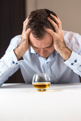 Na kłopoty alkohol. Załamany mężczyzna siedzi przy stole z głową w dłoniach. Szklanka whisky stoi przed nim. - obrazy, fototapety, plakaty