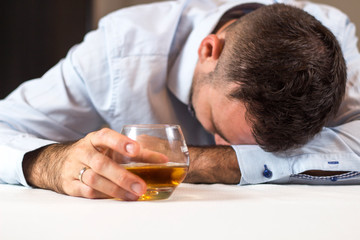 Pijany mężczyzna śpi przy stole z głową opartą o stół. W ręce trzyma szklankę whisky. - obrazy, fototapety, plakaty
