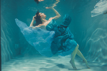 beautiful model girl with long black hair swims underwater in evening blue dress and enjoys...