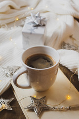Close up view of a white cup of coffee on a book, vintage silver Christmas decorations on a rustic wooden background with a white woolen scarf and a gift or present and Christmas lights 