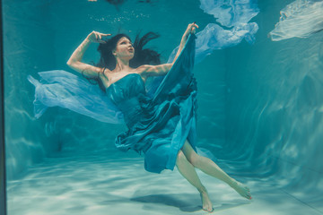 beautiful model girl with long black hair swims underwater in evening blue dress and enjoys...