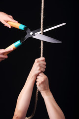 A person hanging from a rope and another one cutting it with big scissors.