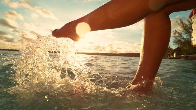 SLOW MOTION, CLOSE UP, LENS FLARE: Unrecognizable playful woman having fun kicking the refreshing ocean water while sitting on the pier on a sunny summer evening in Cook Islands. Girl splashing water.