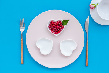 Simple color table setting for celebration with roses, wineberry, pink plates and heart-shaped saucers on blue table background top view