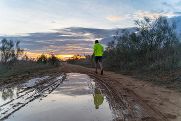 Running in the field at sunset in shorts, with sneakers, along a path with puddles that reflect the sunset.