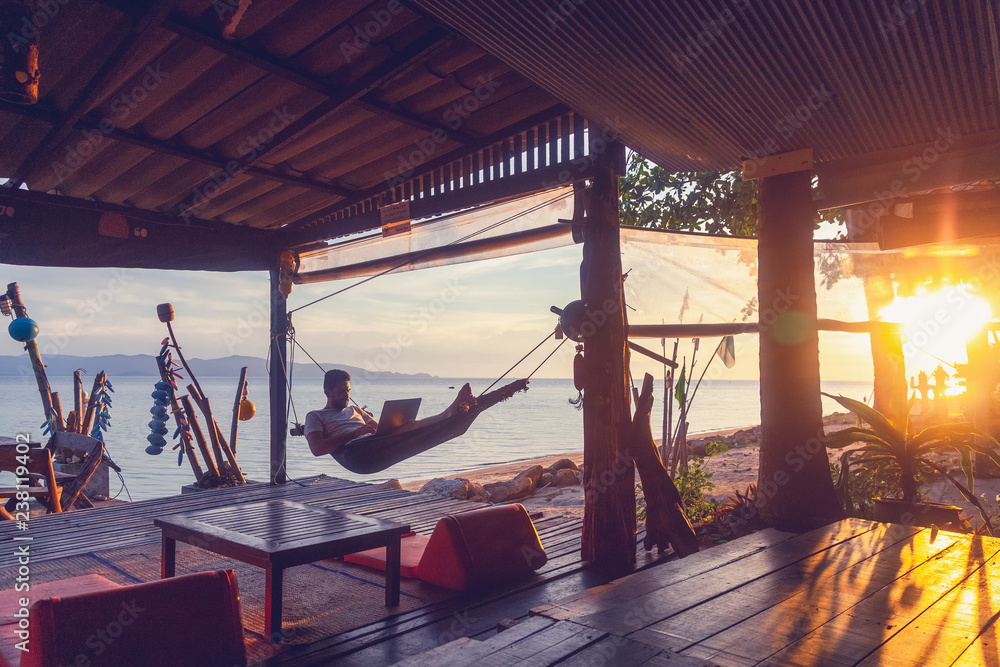 Wall mural young attractive guy a man with a beard in a hammock with a laptop on the background of the sea and 