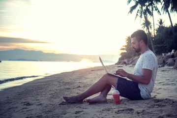 Young attractive guy a man with a beard sitting on the beach with a laptop on the background of the sea and sunset, distant work, freelancer, blogger, vacation and travel