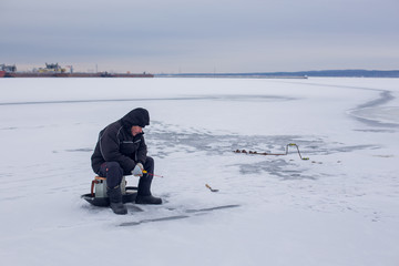 Fototapeta na wymiar Elderly fisherman in dark clothes fishing on winter fishing rod on frozen river on the background of the river port. Winter ice fishing.