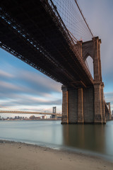 View on Brooklyn Bridge from east river sand beach at sunset with long exposure