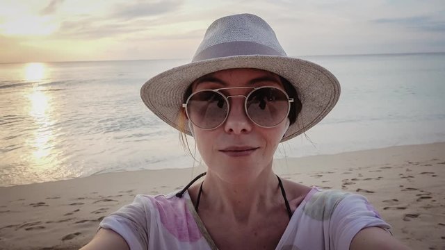 Close up shot of good looking female tourist enjoys free time outdoor near ocean on beach, looks at camera during leisure on sunny summer day, poses for selfie. Happy smiling tourist in tropics sunset