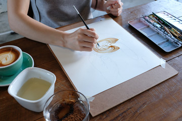 A woman's hand holding a brush painting a coffee cup on white paper with coffee paint on wooden table with props