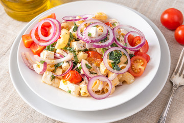 Warm pasta salad, cherry tomatoes, feta cheese and onions on a plate, top view, rustic background