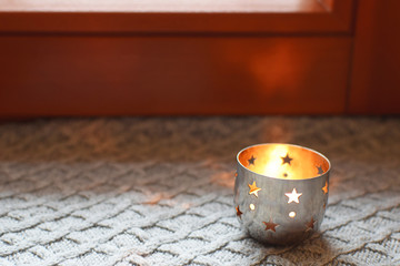 Burning tea candle in small metal holder with star ornament on  grey patterned knitted scarf on brown wooden window sill. Shallow depth of field with focus on candle holder. 