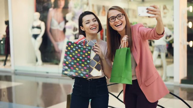 Happy shopaholics are taking selfie with paper bags in shopping mall using smartphone camera holding device and posing, laughing and having fun. Youth lifestyle concept.