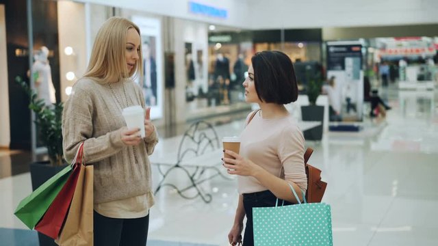 Attractive young women are talking in shopping center holding to-go coffee and paper bags with purchases. Trendy garments on mannequins and glass walls are visible.