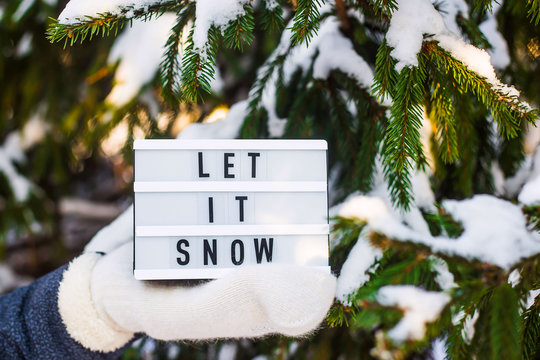 Let it snow it is written on a decorative lamp in the hand of a woman in a white mitten against the background of green spruce branches covered with snow