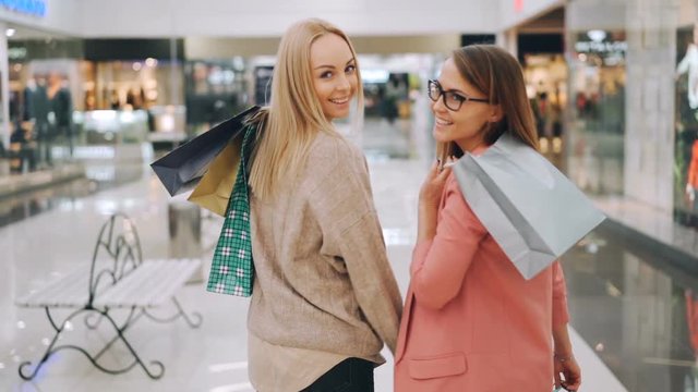 Slow motion of happy young women friends walking together in shopping mall holding bright bags then turning to camera and smiling. Shopaholics and shops concept.