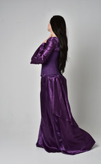 Fototapeta premium full length portrait of beautiful girl with long black hair, wearing purple fantasy medieval gown. standing pose with back to the camera on grey studio background.