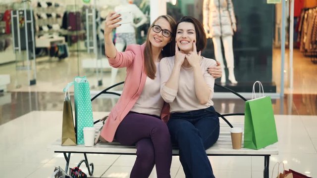 Pretty young women are taking selfie using smartphone camera sitting on bench in shopping mall and having fun. Modern technology, shops and friends concept.