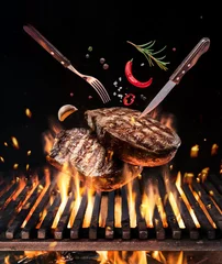 Photo sur Plexiglas Steakhouse Des steaks de bœuf cru avec des légumes et des épices volent au-dessus du feu de barbecue flamboyant.