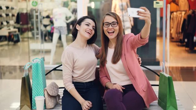 Good-looking young ladies are taking selfie together using smartphone sitting in shopping mall resting and having fun. Girls are hugging and posing with hand gestures.