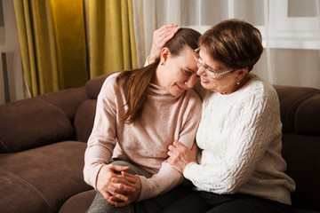 senior mother consoles, comforting sad adult grown daughter sitting at sofa at home. support in two...