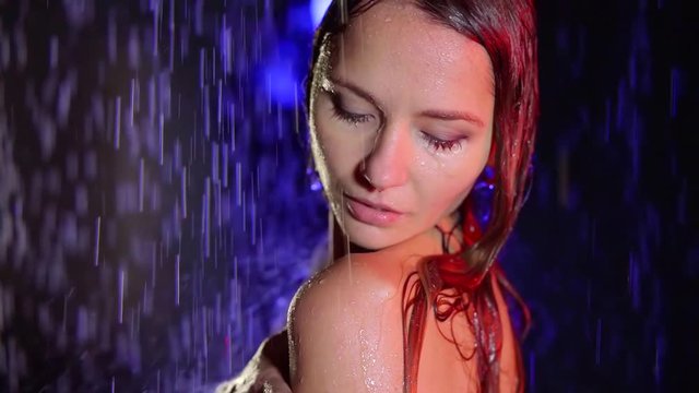 redhead naked woman is standing under rain flows in darkness, touching her shoulder and washing it