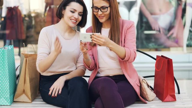 Emotional girls are looking at smartphone screen chatting and laughing sitting on bench in shopping mall with bags and having fun. Modern technology and communication concept.