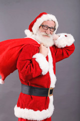 Santa Claus with a white beard wearing glasses and Santa outfit smiling while standing with the bag on the gray background, New Year, Christmas, holidays, souvenirs, gifts, shopping, discounts, shops