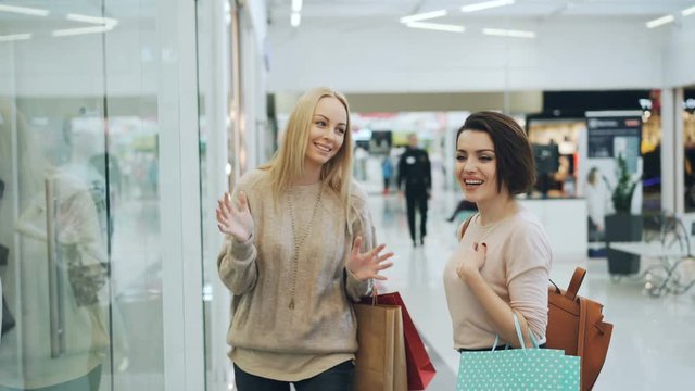 Cute young ladies are discussing lingerie, laughing and chatting in shopping mall standing in front of glass wall pointing at clothing on mannequins. Sale and customers concept.