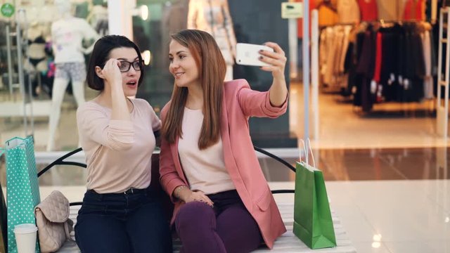 Cheerful female friends are taking selfie with smartphone sitting on bench in shopping mall and posing with glasses making funny faces. Photo, shops and friendship concept.