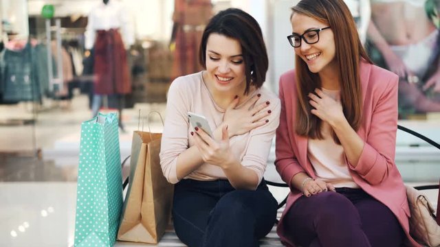 Joyful young women are using smartphone together looking at screen smiling and laughing resting on bench in shopping mall with paper bags visible. Gadgets and fun concept.